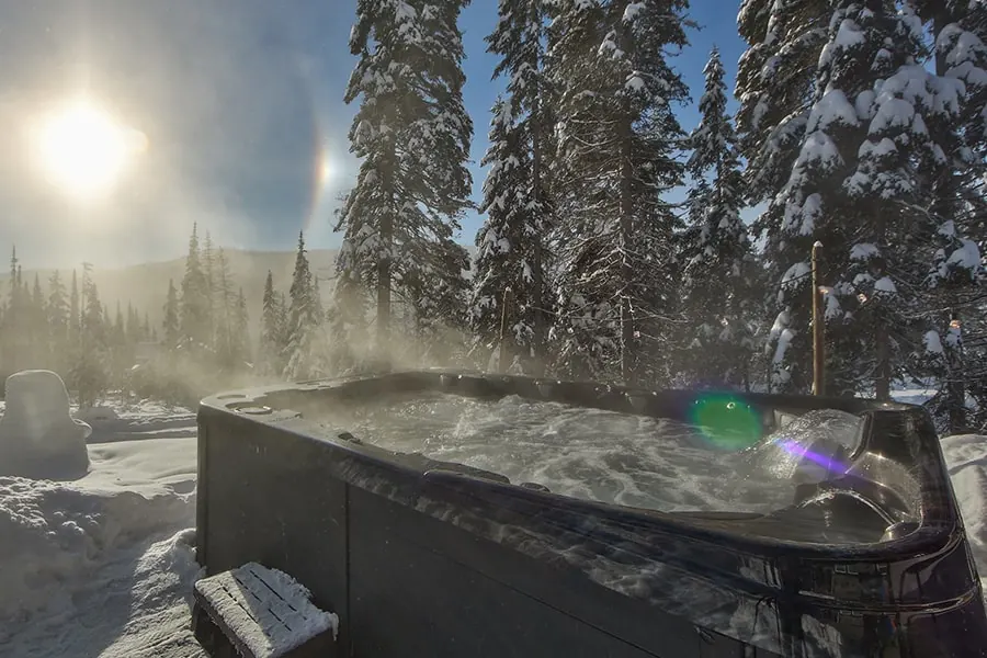 Steam rising off a hot tub in deep snow at the edge of a stand of pines