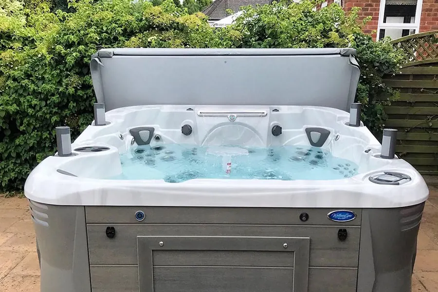 A large hot tub with its cover raised on a paved patio beside a house, backed by mature planting